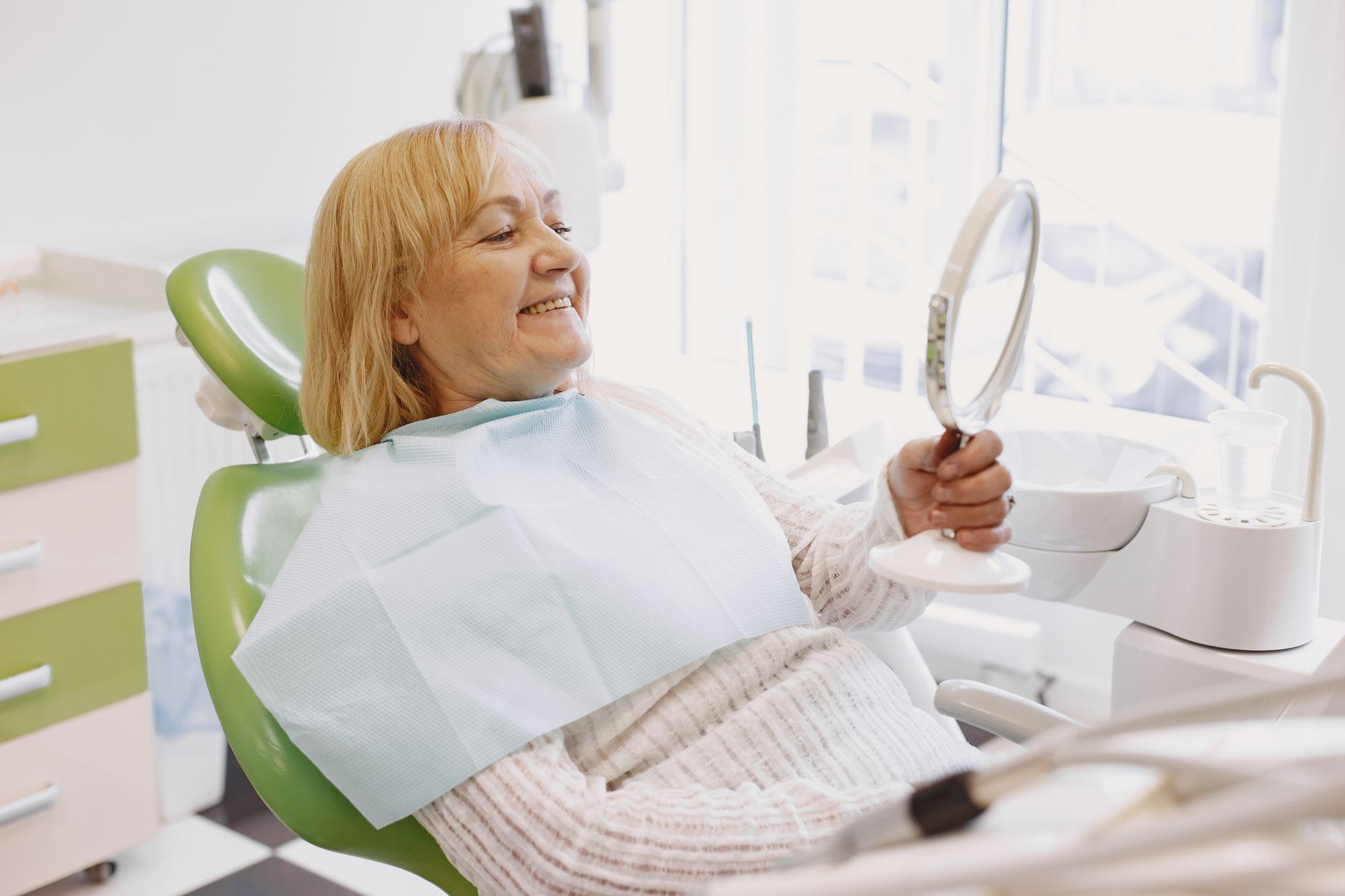 Elderly woman in dentist chair smiling while holding a mirro שיקום הפה והחיוך, ד